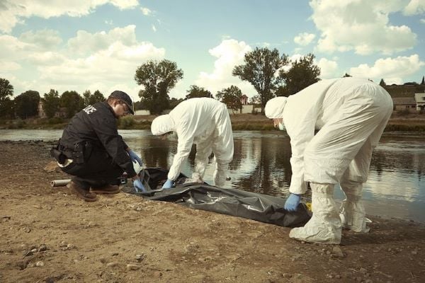 police officer and men in hazmat suits zipping body bag by river